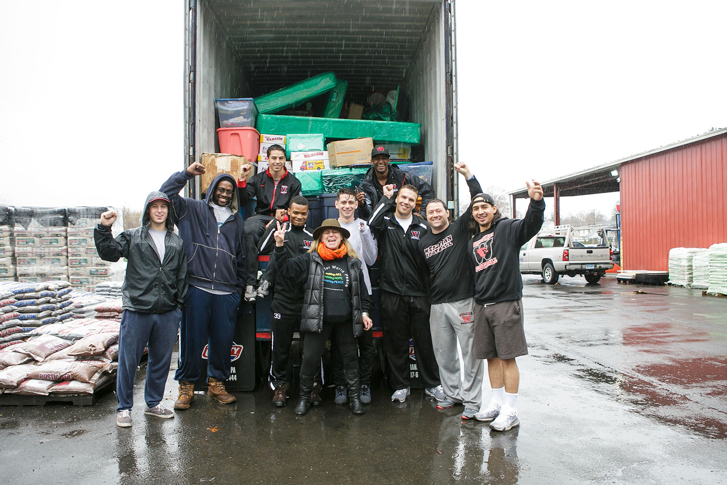 Members of the Wesleyan football team and coach Dan DiCenzo, second from right, celebrate their successful packing service for Artists for World Peace. Founder and executive director Wendy Black-Nasta P’07, center is shipping the equipment to a Tanzanian village where she and others will offer an eye clinic this summer. (Photo by Lucy Guiliano for Artists for World Peace)