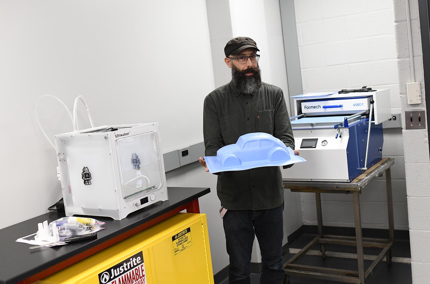 Lopez shows off a vacuum forming tool, which is used to form plastic around a mold and create a permanent object. At left is a 3-D printer.