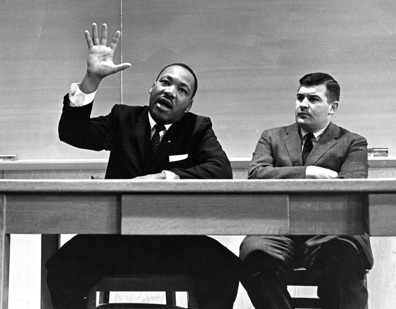 057-Photo-Martin-Luther-King-hand-raised-sitting-with-John-Maguire-at-desk-in-a-classroom-1280x1001.jpg